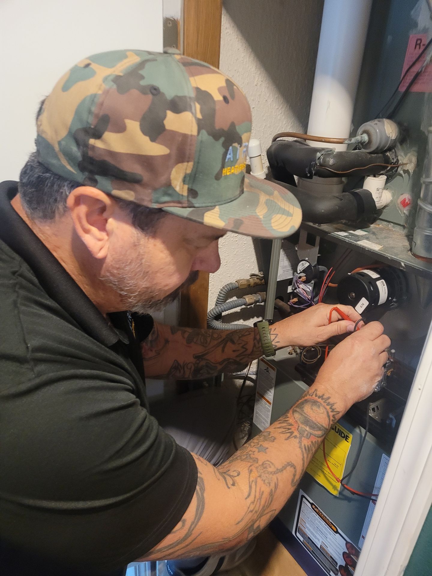 Person in camo cap working on HVAC unit with hands inside open panel.